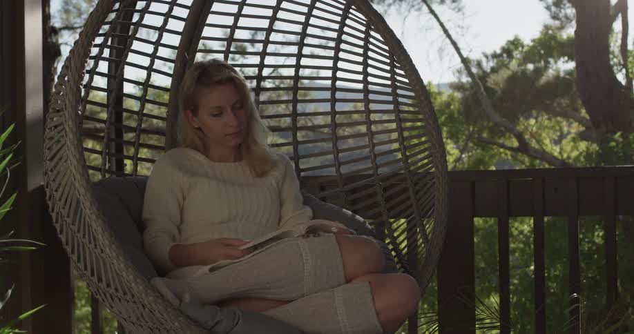 A woman sitting in an egg shaped rattan hanging chair reading a book