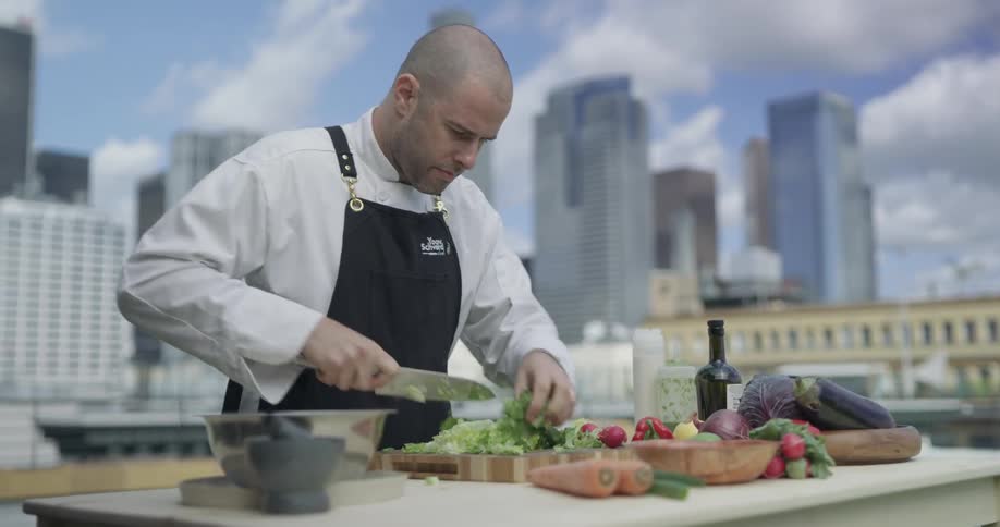 A male chef stand over a table outside cutting lettuce on a cutting board with the city skyline behind him