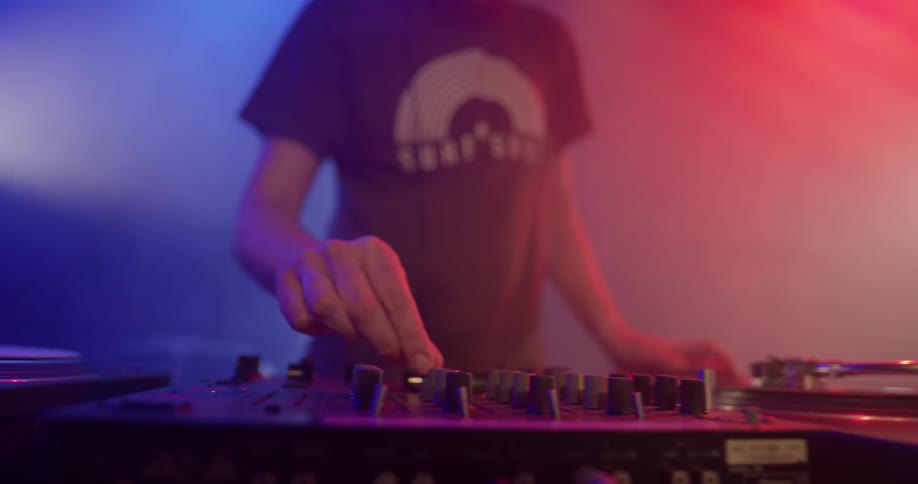 A dj manipulates his equipment as red and purple light shine through smoke behind him