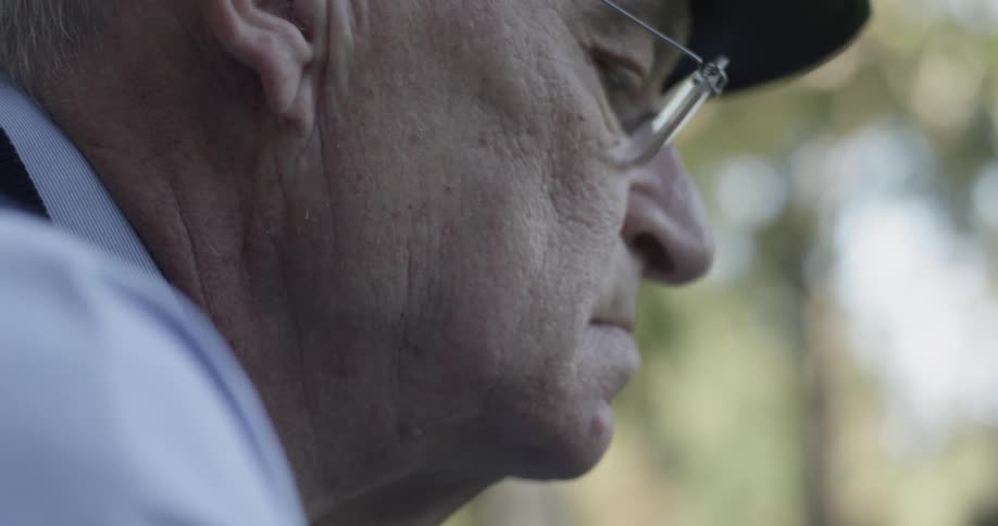 Elderly man wearing glasses and a hat is examining something outside on a sunny day
