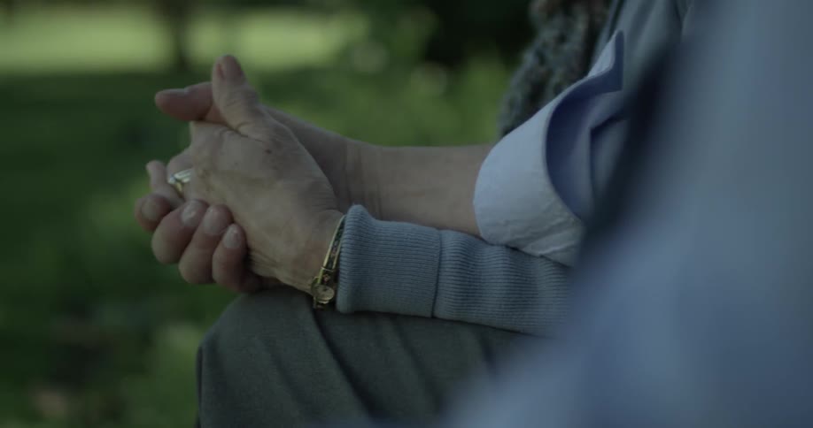 An elderly couple hold hands at the park while sitting on a bench