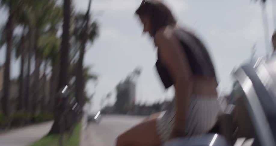 A woman hops out the side of the back seat of a convertible