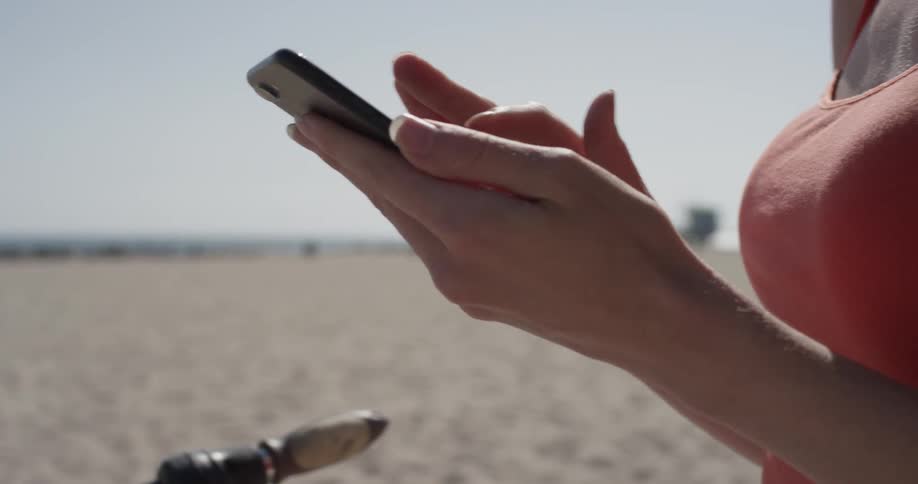 A woman swipes through her phone standing on the beach
