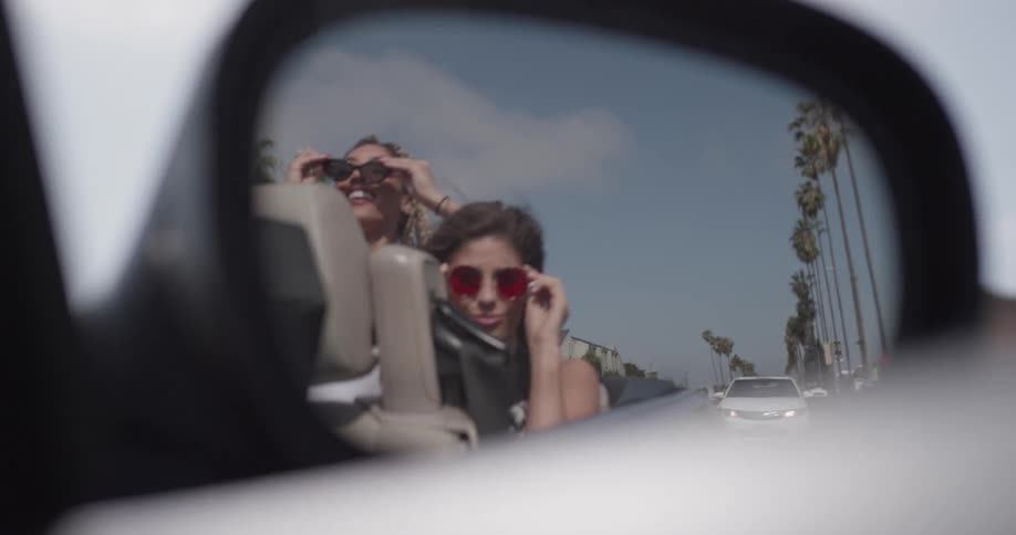 Two women's reflections in a car's side mirror dance to music and put on sunglasses while the car is moving