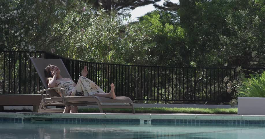 Woman sits on a lounge chair by a pool talking on the phone