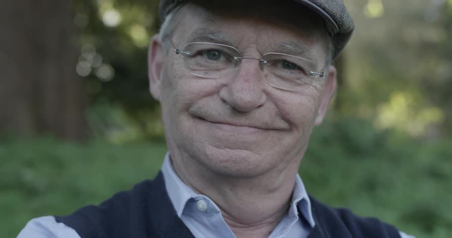 A man smiles and is surprised by the camera wearing glasses and a hat with a sweater vest