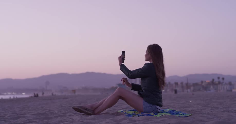 A woman sitting on a towel at the beach taking video of the ocean and chatting on her phone