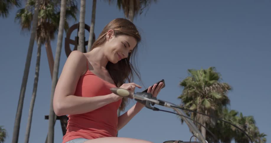 A woman straddles a bike as she looks at a phone near palm trees