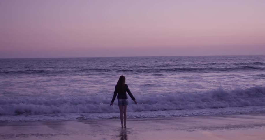 Woman walks towards the ocean at sunset