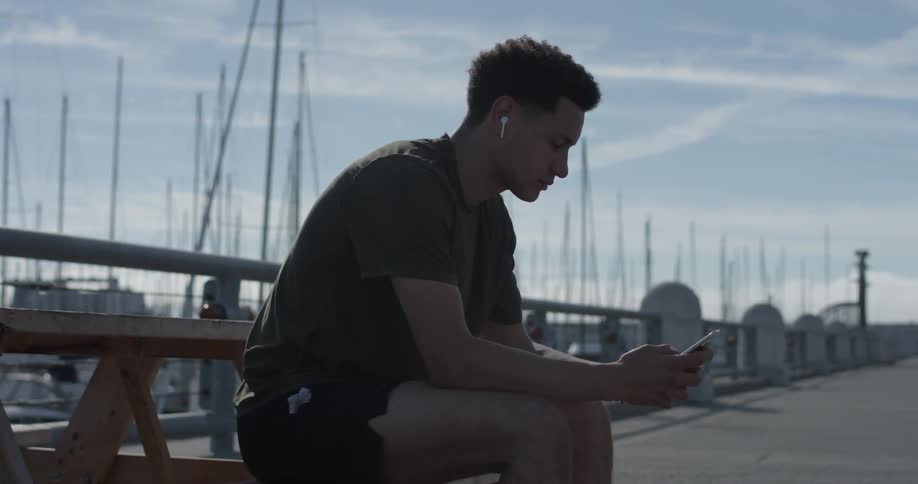 A man sits on a picnic table on a pier and listens to music from his phone through earbuds