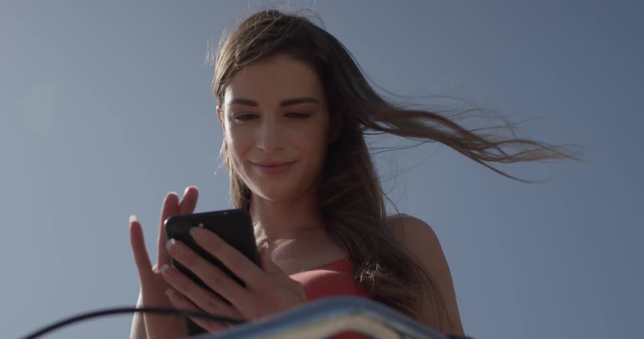 A woman pulls a phone out of her back pocket and smiles at it as she straddles a bike