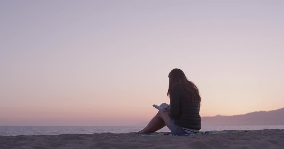 Woman sits on the beach at sunrise looking at a book