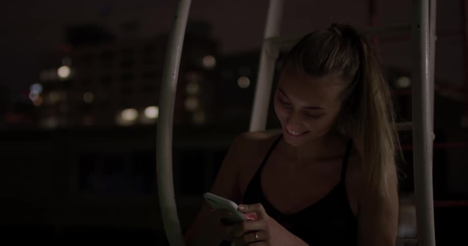 A woman smiles as she texts on her phone while sitting on fire escape steps in the city