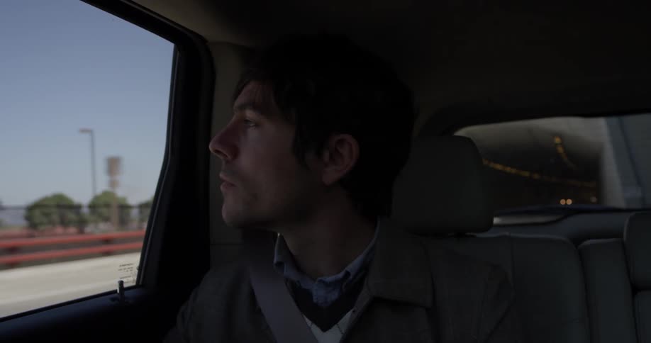 A man sits in the backseat of a car looking out the window at the city on a bright sunny day
