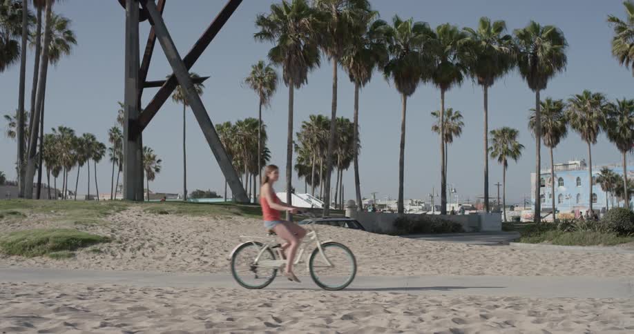 Woman pedals her bike on a sidewalk past the sandy beach