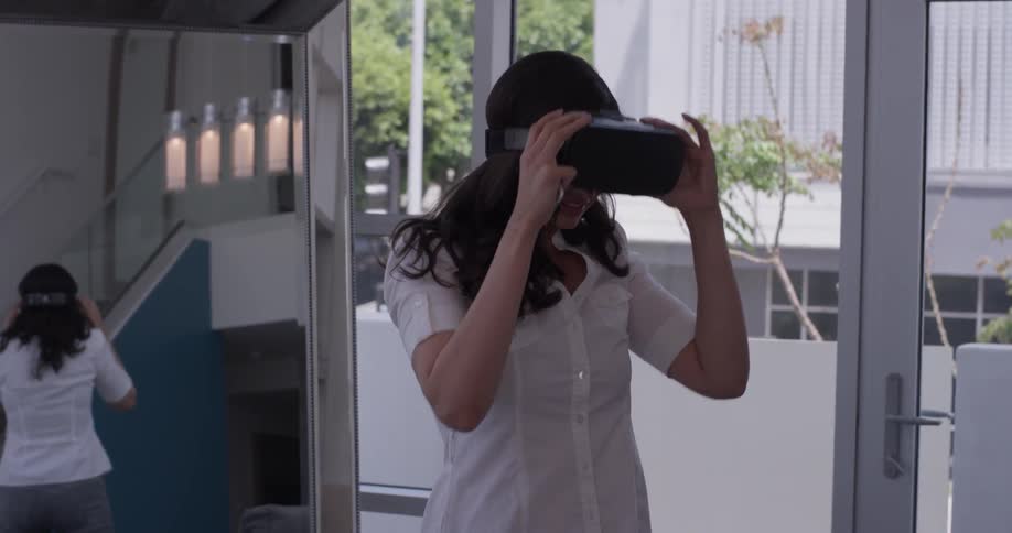 A woman stands in a room wearing virtual reality goggles with a remote controller in her hands as she takes off the goggles and inspects them