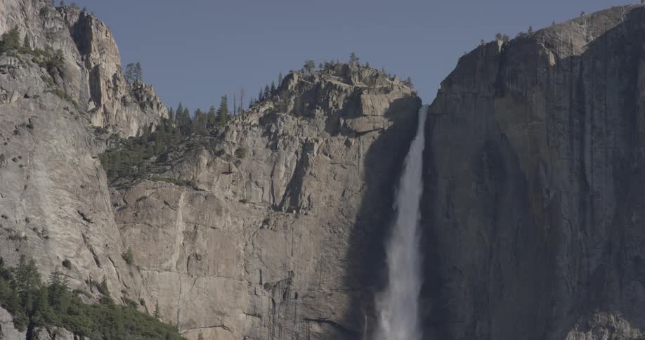 A waterfall has carved out a slit in the mountain with trees on it and a blue sky background on a sunny day