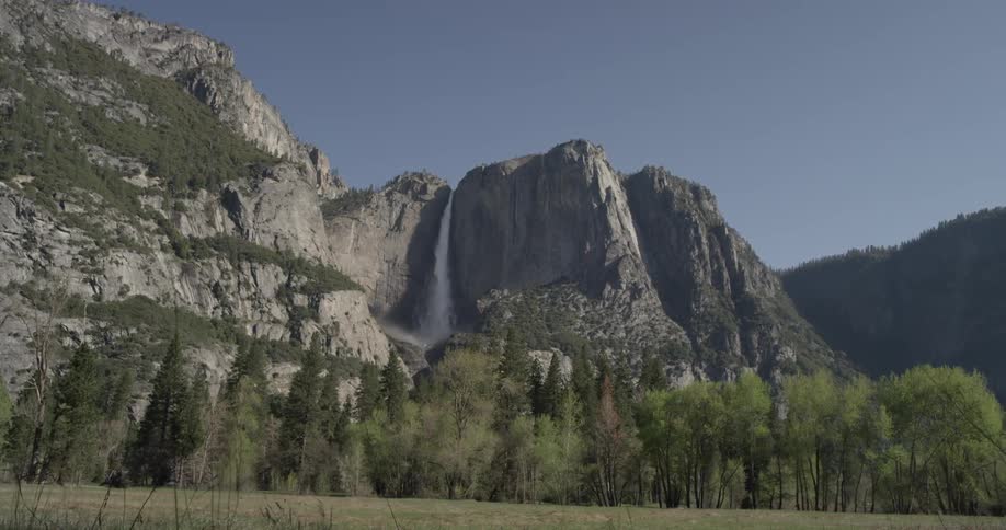 Water pours off the side of a mountain into a valley with grassy field and plenty of green trees