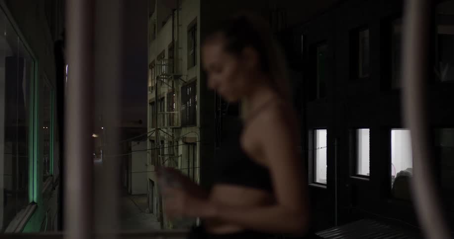 A woman stands on a fire escape and drinks a bottle of water at night