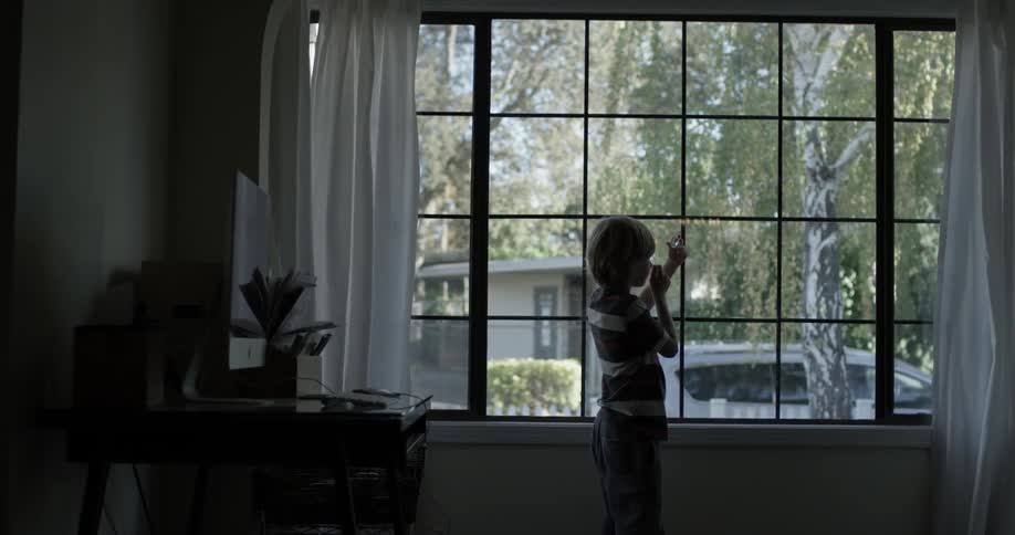 Child looks at an object in one hand with a magnifying glass in the other in front of a window in his living room
