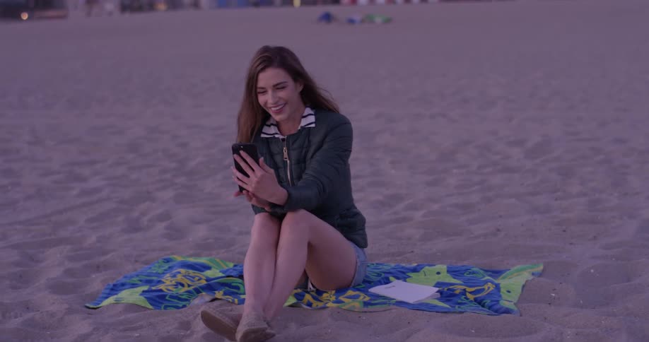 A woman smiles at her phone as she sits on the beach on a beach towel