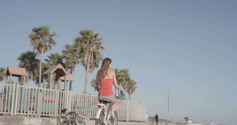 A woman rides her bike past a fenced in jungle gym on the beach