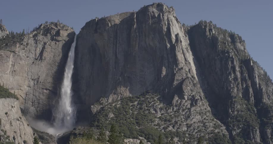 A lot of water pours down the side of a mountain and hitting many rocks below creating mist and humidity for the plants and trees below