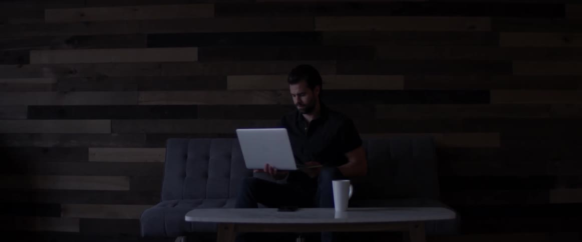 A man sits on a couch and uses his phone then sets it down and puts a laptop on his lap and grabs a cup of coffee