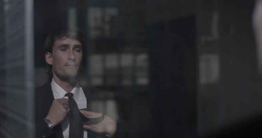 A man stands in front of a large window overlooking a city and adjusts his tie
