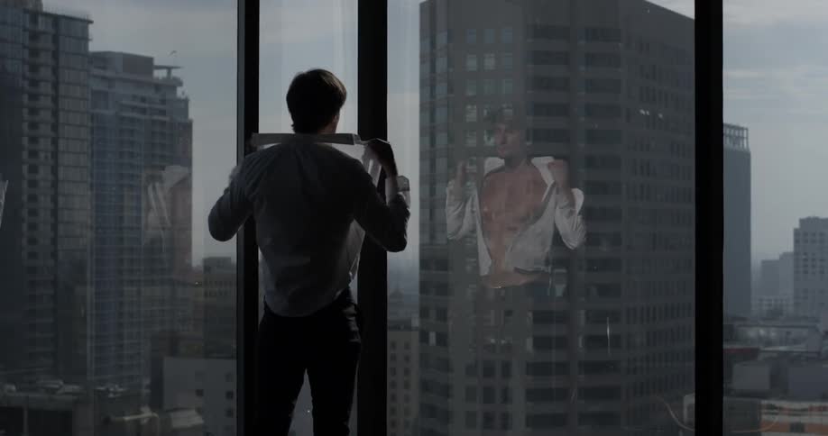 A man puts on a white shirt as he looks at a cities skyline that's under an overcast sky