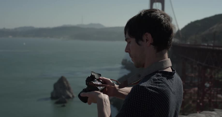A man lifts a camera to his eye and takes photos towards a body of water