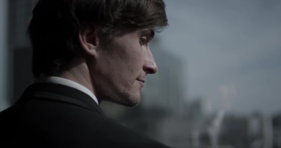 A man stands in front of a window wearing a suit while sun beams through the glass on to his face as he looks out at the city