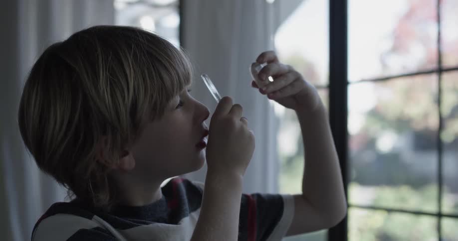 A boy looks at a glass oval through the magnifying glass and stands in the living room in front of a window with sunlight coming in