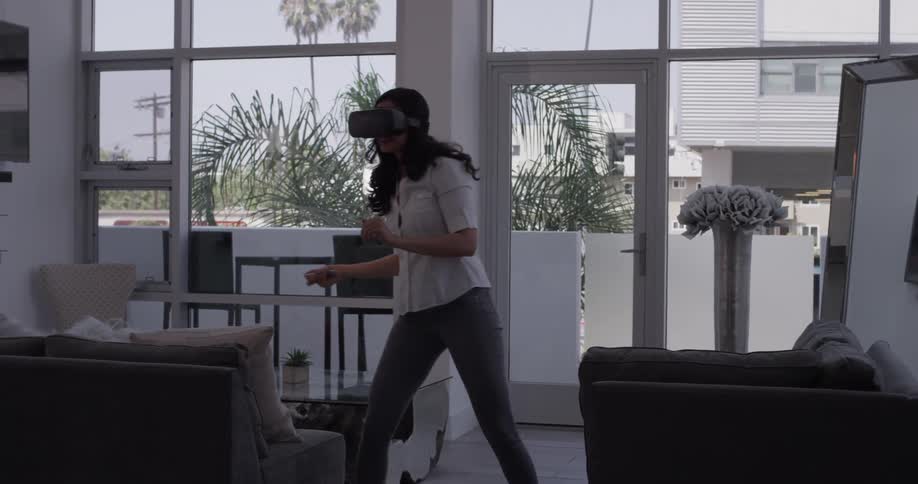 A woman wearing virtual reality goggles is playing a video game in her living room while standing