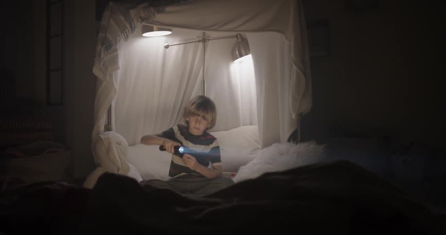 A little boy shines a flashlight from his makeshift tent he made with sheets in his bedroom