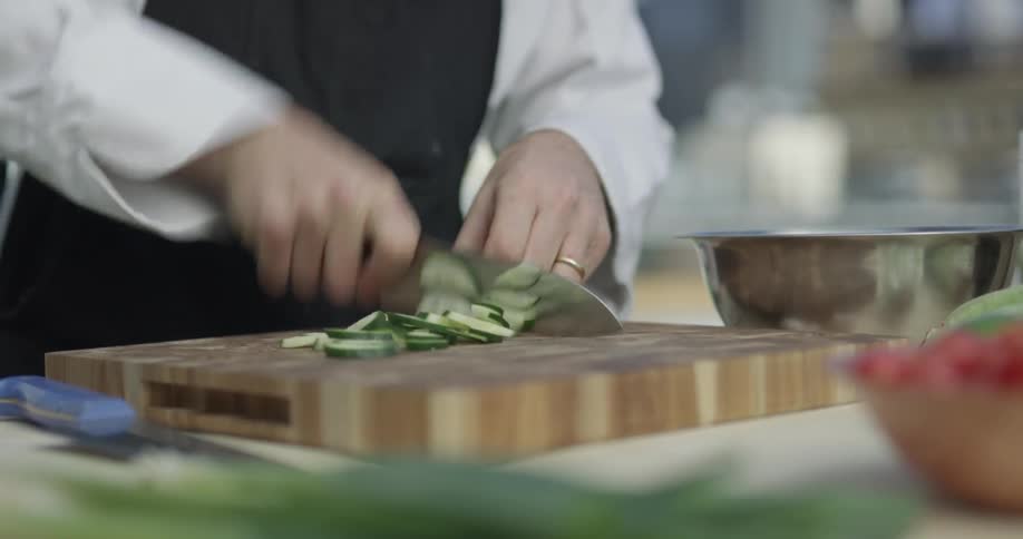 A chef cuts up one half of a cucumber on a wooden cutting board on the kitchen counter top