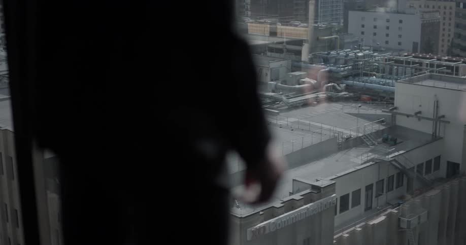 A person in a suit is standing in a room overlooking a city through a very large window in the wall