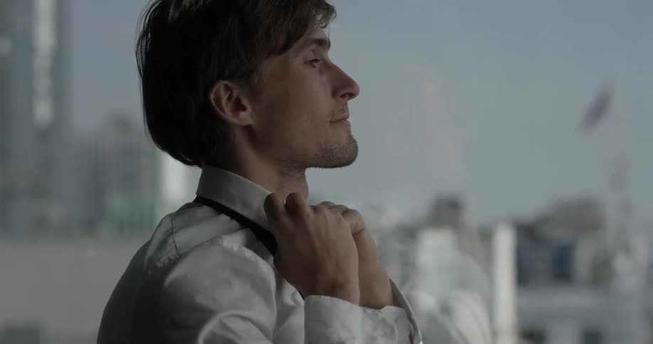 A man adjusts his collar and puts on a tie while looking out at the city through the tall windows