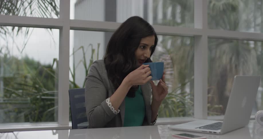 A woman works on her laptop and drinks hot coffee out of a mug