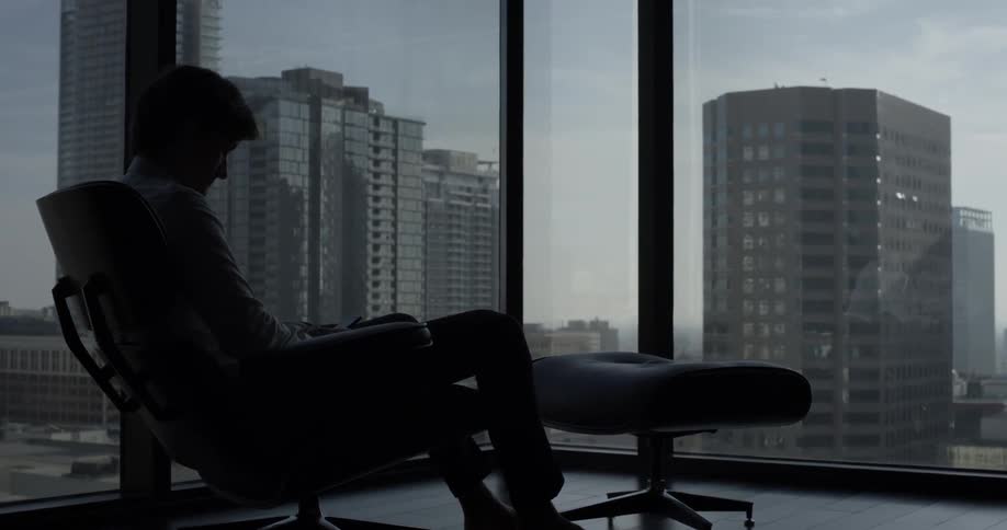 A man sits in the corner of an office in a chair overlooking the city through large tall windows
