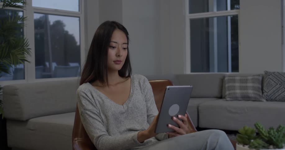 An asian woman sits in a chair indoors and works on her tablet