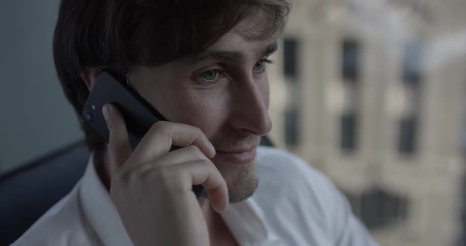 Man sits in a chair in the office holding a phone to his ear and talks on it with large windows in the background overlooking the city