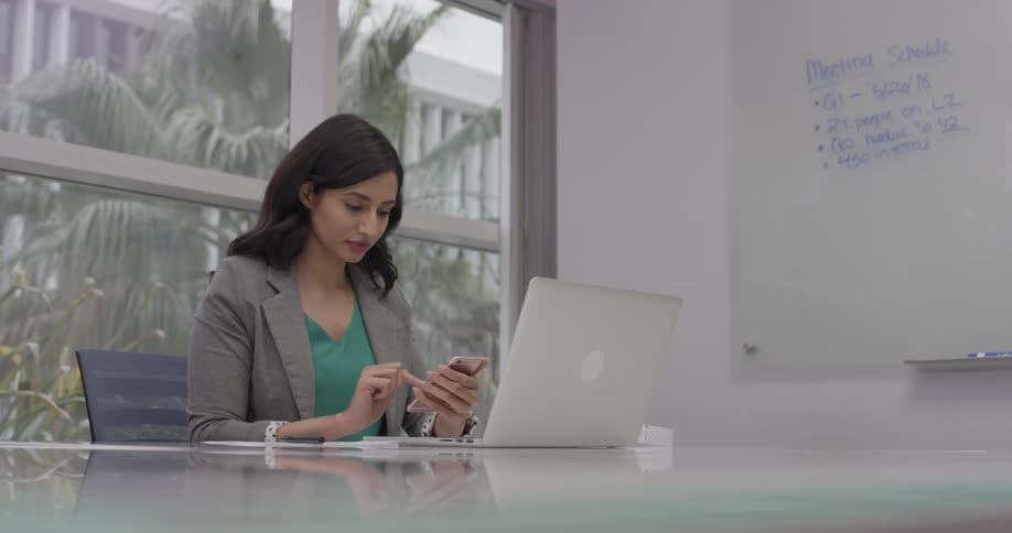 Woman working in office on a laptop and cellphone in front of a wall of windows