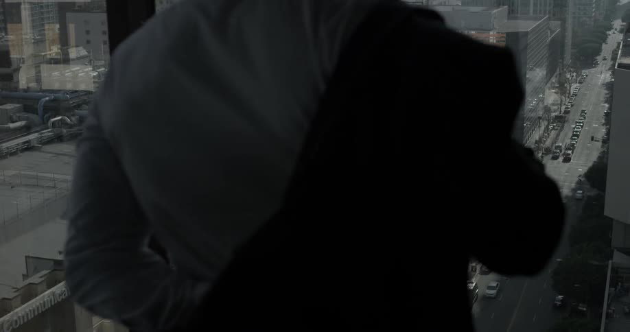 A man throws on a black jacket over his white shirt in the corner of room with large windows overlooking the city