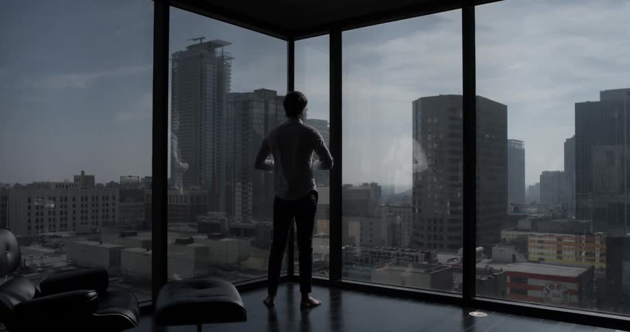 A man shrugs on his shirt and buttons it as he looks at the skyline through his high rise windows