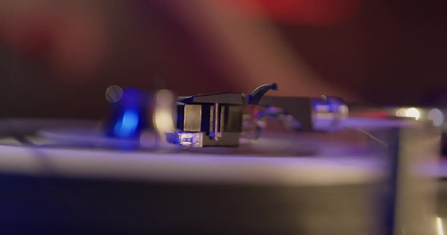 A record is played on a record player by a dj moving his hands in the background with red and blue lights shining