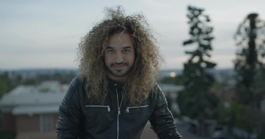 A man is smiling outside as the sun sets and he stands on top of a roof top out in the open of a cloudy day