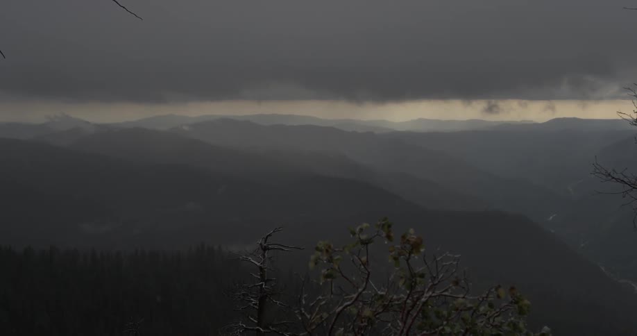 Rain falls on a cloudy dimly lit day over a landscape of mountains covered with trees