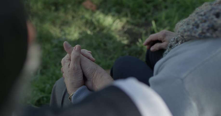 A man grabs the hand of an elderly woman next to him on a park bench and their thumbs mingle
