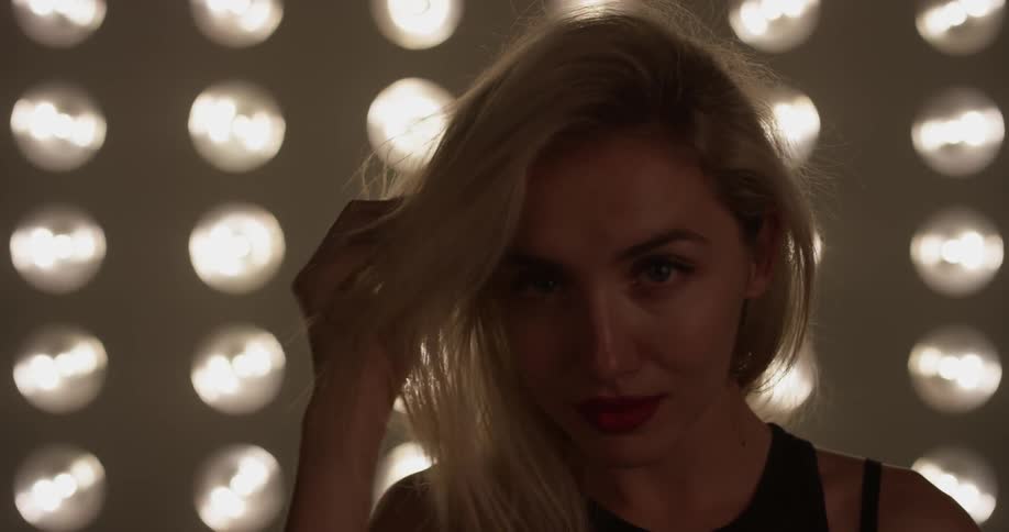 A woman stands in front of large lights and touches her hair